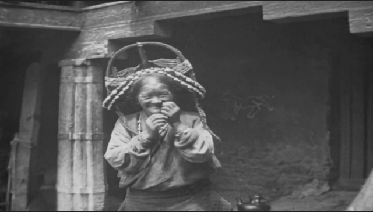 An elderly woman stands near a pillar, smiling and holding her hands near her face. She wears an ornate headdress and a beaded necklace.
