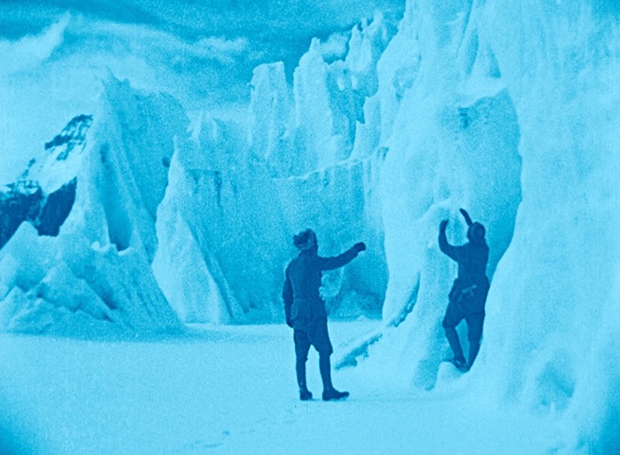 In a blue saturated frame, two men examine icy cliffs that reach only around two stories high. The snowy ground is strangely flat.